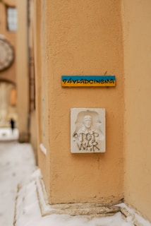 A corner of an urban building with textured beige walls. There is a small sign with blue and yellow coloring, displaying the text '24VLADCINEMA.' Below the sign, a white relief sculpture with a face and the text 'STOP WAR' is visible. Snow is accumulated at the base of the building, indicating a cold climate.