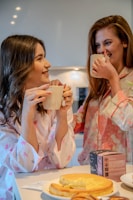 Two women are enjoying tea and laughing together in a kitchen while wearing floral pajamas. In the foreground, there is a table with a slice of cheesecake and a box of tea.