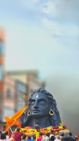 A large black statue of a deity with closed eyes, adorned with large earrings and a prominent teardrop-shaped marking on the forehead, surrounded by colorful garlands. Several people are gathered around the statue, some holding orange flags. The background features blurred buildings and a clear blue sky.