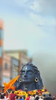 A large black statue of a deity with closed eyes, adorned with large earrings and a prominent teardrop-shaped marking on the forehead, surrounded by colorful garlands. Several people are gathered around the statue, some holding orange flags. The background features blurred buildings and a clear blue sky.