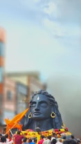 A large black statue of a deity with closed eyes, adorned with large earrings and a prominent teardrop-shaped marking on the forehead, surrounded by colorful garlands. Several people are gathered around the statue, some holding orange flags. The background features blurred buildings and a clear blue sky.