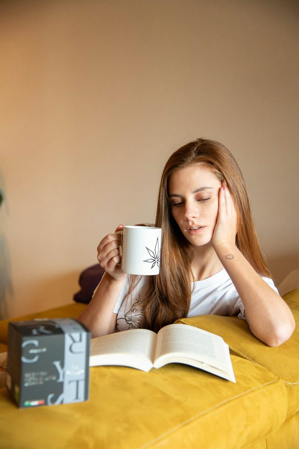 Eine Frau sitzt auf einer Couch und hält ein Buch und eine Tasse Kaffee in der Hand