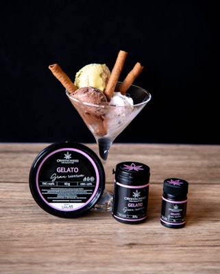 A glass cup filled with three scoops of gelato in different flavors, accompanied by three wafer sticks. Beside the cup are three containers labeled 'Crystalweed Gelato Gran Riserva', indicating the product's THC and CBD content.