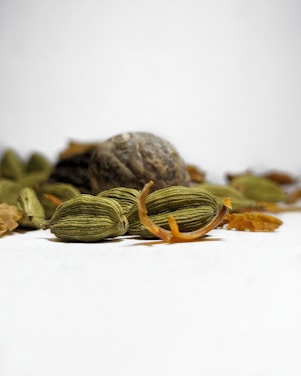 Close-up view of cardamom pods and nutmeg placed on a plain surface with a soft focus background. The cardamom pods are green and elongated with some scattered brown peelings around them, while the nutmeg appears round and textured.