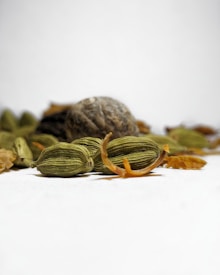 Close-up view of cardamom pods and nutmeg placed on a plain surface with a soft focus background. The cardamom pods are green and elongated with some scattered brown peelings around them, while the nutmeg appears round and textured.