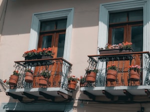 Two balconies with ornate wrought iron railings adorned with vibrant pots of red and pink flowers. The wooden window frames are distinguished with a light blue trim, creating a warm and serene architectural detail.