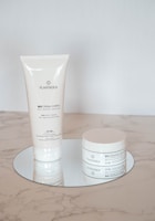 Two skincare products with a minimalistic white design are placed on a reflective round surface. One is a tall, slender tube, and the other is a round jar. Both are labeled with the brand name 'PLANTADEA' and appear to be for dry and delicate skin, containing 1% CBD.