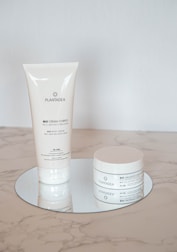 Two skincare products with a minimalistic white design are placed on a reflective round surface. One is a tall, slender tube, and the other is a round jar. Both are labeled with the brand name 'PLANTADEA' and appear to be for dry and delicate skin, containing 1% CBD.