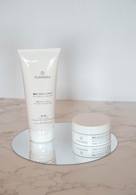 Two skincare products with a minimalistic white design are placed on a reflective round surface. One is a tall, slender tube, and the other is a round jar. Both are labeled with the brand name 'PLANTADEA' and appear to be for dry and delicate skin, containing 1% CBD.