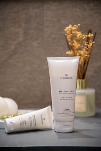A tube of Plantadea Bio Crema Corpo, a body cream for dry and delicate skin, standing upright on a surface. Another tube of the same cream lies next to it. The background includes a small vase filled with dried flowers and another container, creating a calm and natural setting.