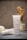 A tube of Plantadea Bio Crema Corpo, a body cream for dry and delicate skin, standing upright on a surface. Another tube of the same cream lies next to it. The background includes a small vase filled with dried flowers and another container, creating a calm and natural setting.