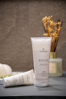 A tube of Plantadea Bio Crema Corpo, a body cream for dry and delicate skin, standing upright on a surface. Another tube of the same cream lies next to it. The background includes a small vase filled with dried flowers and another container, creating a calm and natural setting.
