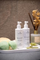 A neatly arranged bathroom setup features two bottles of skincare products labeled with the brand name 'PlantaDea' placed on a white tray. Nearby, two green sponge-like items, a yellow pumice stone, and a jade roller are artfully arranged. In the background, dried flowers are held in a diffuser bottle.