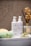 A neatly arranged bathroom setup features two bottles of skincare products labeled with the brand name 'PlantaDea' placed on a white tray. Nearby, two green sponge-like items, a yellow pumice stone, and a jade roller are artfully arranged. In the background, dried flowers are held in a diffuser bottle.
