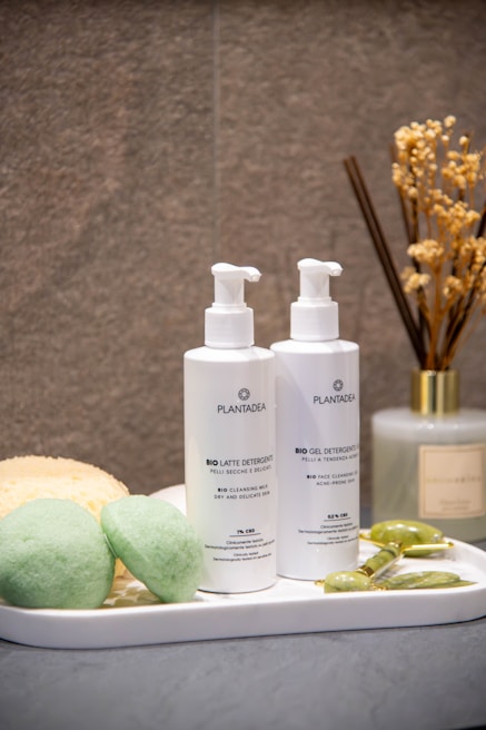 A neatly arranged bathroom setup features two bottles of skincare products labeled with the brand name 'PlantaDea' placed on a white tray. Nearby, two green sponge-like items, a yellow pumice stone, and a jade roller are artfully arranged. In the background, dried flowers are held in a diffuser bottle.