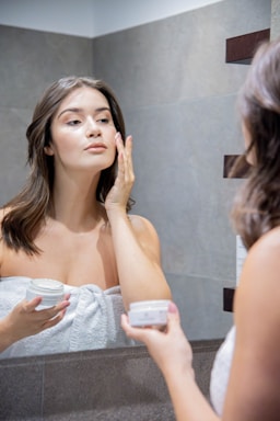 A woman stands in front of a bathroom mirror applying cream to her face. She is wrapped in a white towel, holding a jar of skincare cream in one hand while gently touching her face with the other. The setting is a modern bathroom with tiled walls.