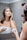 A woman stands in front of a bathroom mirror applying cream to her face. She is wrapped in a white towel, holding a jar of skincare cream in one hand while gently touching her face with the other. The setting is a modern bathroom with tiled walls.