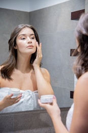 A woman stands in front of a bathroom mirror applying cream to her face. She is wrapped in a white towel, holding a jar of skincare cream in one hand while gently touching her face with the other. The setting is a modern bathroom with tiled walls.