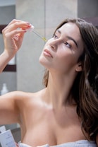 A woman is applying a skincare serum to her face using a dropper. Her eyes are gazing upwards as she delicately holds the dropper above her skin. She is wrapped in a towel, suggesting a beauty or skincare routine in a bathroom setting.