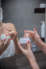 Hands with red nail polish are holding and applying a face cream in front of a mirror. The person appears to be in a bathroom setting with a towel wrapped around their body.
