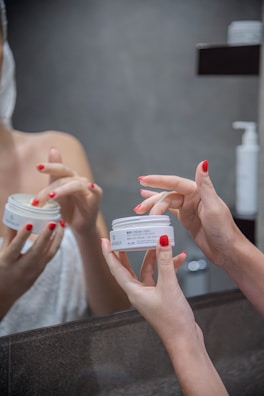 Hands with red nail polish are holding and applying a face cream in front of a mirror. The person appears to be in a bathroom setting with a towel wrapped around their body.