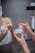 Hands with red nail polish are holding and applying a face cream in front of a mirror. The person appears to be in a bathroom setting with a towel wrapped around their body.