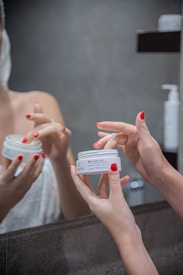 Hands with red nail polish are holding and applying a face cream in front of a mirror. The person appears to be in a bathroom setting with a towel wrapped around their body.
