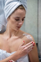 A person wrapped in a towel with a towel turban on their head applies lotion to their shoulder. The setting appears to be a bathroom with neutral-colored walls, and the person's nails are painted red.