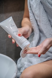 A person is applying cream from a tube onto their hand. The person is draped in a white towel and has red painted nails. The scene appears to be in a bathroom setting.
