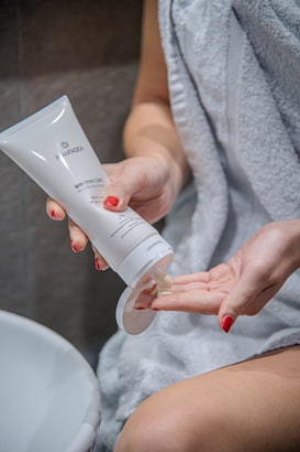 A person is applying cream from a tube onto their hand. The person is draped in a white towel and has red painted nails. The scene appears to be in a bathroom setting.