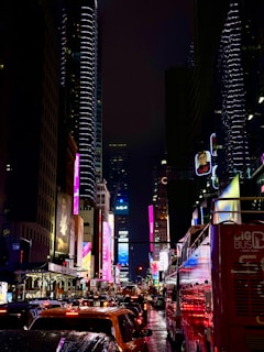 A busy city street at night with heavy traffic, surrounded by tall skyscrapers adorned with bright and colorful billboards and neon lights. The street is wet, reflecting the lights, while various advertisements and signs illuminate the scene. A large red bus is visible to the right side, with multiple cars lined up in traffic.