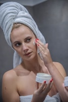 A person with a towel wrapped around their head applies a cream to their face. The individual has red nail polish and holds a jar of cream, focusing on their skincare routine in a bathroom setting.
