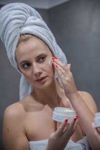 A person with a towel wrapped around their head applies a cream to their face. The individual has red nail polish and holds a jar of cream, focusing on their skincare routine in a bathroom setting.