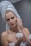 A person with a towel wrapped around their head applies a cream to their face. The individual has red nail polish and holds a jar of cream, focusing on their skincare routine in a bathroom setting.