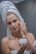 A person with a towel wrapped around their head applies a cream to their face. The individual has red nail polish and holds a jar of cream, focusing on their skincare routine in a bathroom setting.