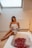 A woman wrapped in a towel is sitting on the edge of a bathtub filled with water and red rose petals. The setting is softly lit, creating a relaxing atmosphere. A candle is placed near the tub and a skylight above provides natural light.