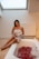 A woman wrapped in a towel is sitting on the edge of a bathtub filled with water and red rose petals. The setting is softly lit, creating a relaxing atmosphere. A candle is placed near the tub and a skylight above provides natural light.