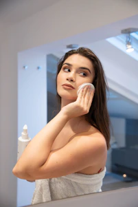 A cheerful woman happily applying skincare product in a bright, cozy bathroom.