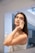 A woman stands in front of a mirror in a bathroom, gently applying a product to her face with a cotton pad. She is holding a bottle in one hand, which appears to be a skincare product. Her hair is down, and she is wrapped in a towel.