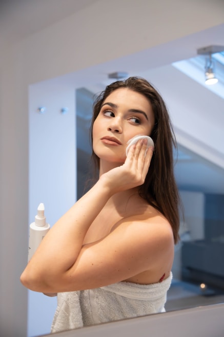 A woman stands in front of a mirror in a bathroom, gently applying a product to her face with a cotton pad. She is holding a bottle in one hand, which appears to be a skincare product. Her hair is down, and she is wrapped in a towel.