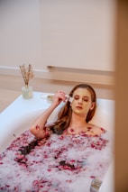 A person is relaxing in a bathtub filled with water and rose petals. The person has a facial mask applied and is using a brush to apply or adjust the mask. The setting is peaceful with a neutral-colored bathroom and a small vase with dry flowers on the side.