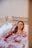 A person is relaxing in a bathtub filled with water and rose petals. The person has a facial mask applied and is using a brush to apply or adjust the mask. The setting is peaceful with a neutral-colored bathroom and a small vase with dry flowers on the side.