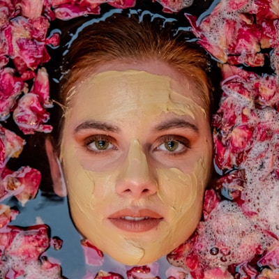 A woman's face emerges from a surface surrounded by vibrant pink rose petals, with a creamy facial mask applied to her skin.