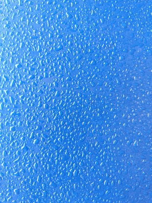 Multiple droplets are scattered across a smooth blue surface, creating a textured and shiny appearance. The droplets vary in size and shape, reflecting light.