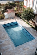 An empty rectangular swimming pool with blue tiles is surrounded by a paved area. The pool has some fallen leaves on its surface. Nearby is a small structure with a red roof and a white door, with a garden area filled with trees and bushes in the background.