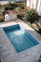 An empty rectangular swimming pool with blue tiles is surrounded by a paved area. The pool has some fallen leaves on its surface. Nearby is a small structure with a red roof and a white door, with a garden area filled with trees and bushes in the background.