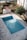 An empty rectangular swimming pool with blue tiles is surrounded by a paved area. The pool has some fallen leaves on its surface. Nearby is a small structure with a red roof and a white door, with a garden area filled with trees and bushes in the background.
