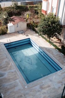 An empty rectangular swimming pool with blue tiles is surrounded by a paved area. The pool has some fallen leaves on its surface. Nearby is a small structure with a red roof and a white door, with a garden area filled with trees and bushes in the background.