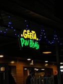 A restaurant sign reads 'Geeta Pav Bhaji' in bright yellow and green letters. Blue decorative lights are strung around the sign, adding a festive appearance. The background shows a wooden exterior with warm lighting overhead, suggesting an inviting atmosphere. People are partially visible, indicating activity inside the establishment.