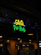 A restaurant sign reads 'Geeta Pav Bhaji' in bright yellow and green letters. Blue decorative lights are strung around the sign, adding a festive appearance. The background shows a wooden exterior with warm lighting overhead, suggesting an inviting atmosphere. People are partially visible, indicating activity inside the establishment.